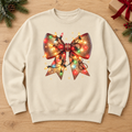 An off-white crewneck sweatshirt laid flat on a light wood background, decorated with a large Christmas bow graphic filled with glowing multicolored holiday lights. Pine branches, a gift box, and a red ribbon are visible around the edges as seasonal props.