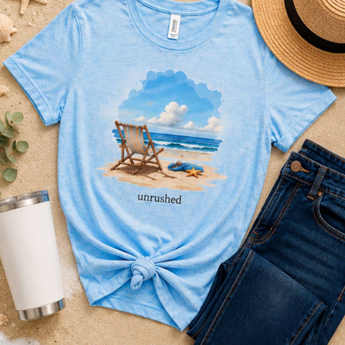 Blue t-shirt with beach scene and 'unrushed' text, placed on sand with accessories.