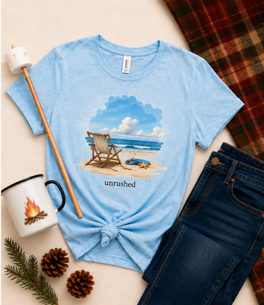 Light blue t-shirt with beach graphic and 'unrushed' text, placed on a surface with a mug, plaid fabric, and pine branches.