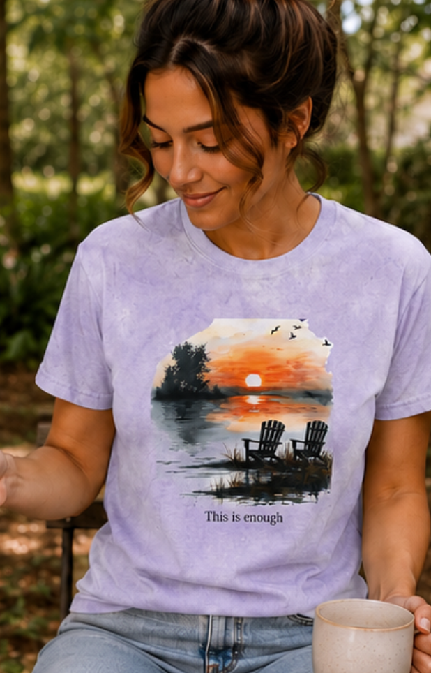 Woman wearing a t-shirt with a sunset design and 'This is enough' text, sitting outdoors.
