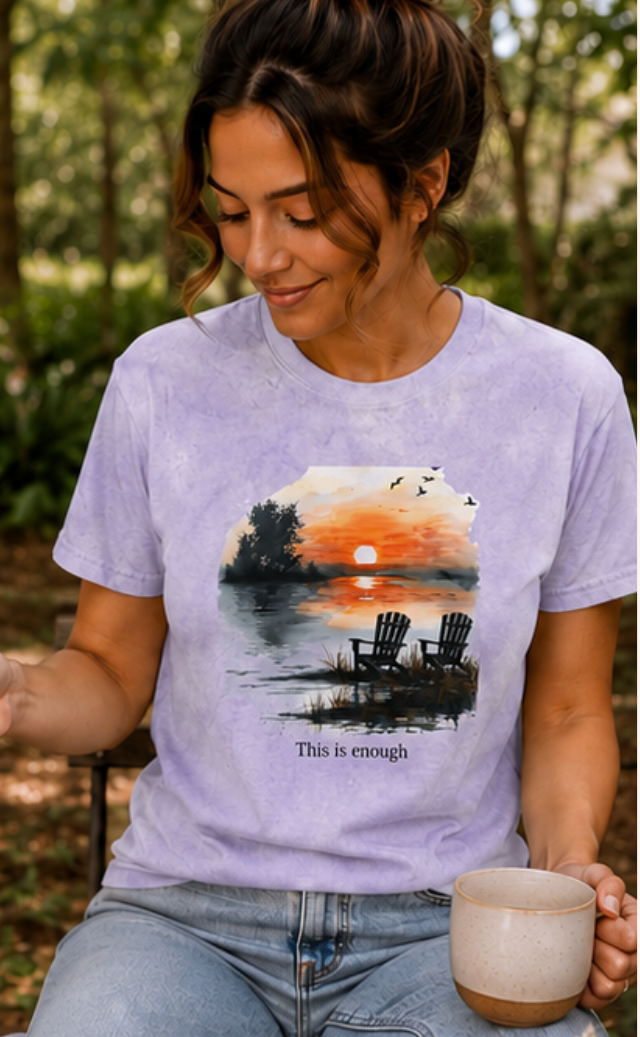 Woman wearing a t-shirt with a sunset design and 'This is enough' text, sitting outdoors.