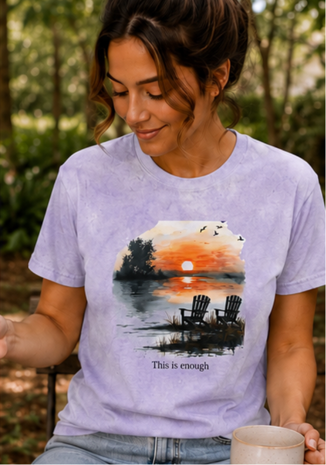 Woman wearing a t-shirt with a sunset design and 'This is enough' text, sitting outdoors.