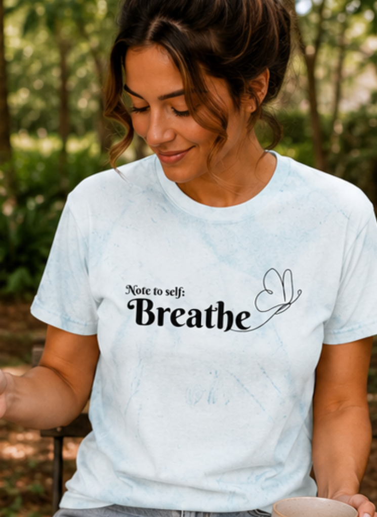 Woman wearing a white t-shirt with 'Note to self: Breathe' text, sitting outdoors.